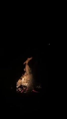 A bonfire burns in the darkness, casting an orange glow on the surrounding logs and smoke. Silhouettes of at least two people are visible on the left side of the frame, facing the fire. The sky above is black, with a few faint stars or distant lights visible. The scene is still and quiet, with the crackling of the fire being the only apparent sound.