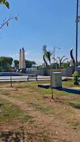 The video shows an outdoor park area on a sunny day with a clear blue sky. In the background, there are several architectural elements, including two tall, white, pointed structures. To the right of these structures, there are dark, curved sculptures. A light pole with multiple lights stands prominently in the center right of the frame. Further to the right, a building with orange-barred windows is visible, with a blue and red tent set up in front of it. The foreground is a grassy area with dry patches of dirt and scattered small plants. There are also several concrete bases with yellow tops scattered across the grassy area, each with a young tree growing from it. A metal box on a concrete base is positioned in the center left of the frame. The camera pans slightly to the right, revealing a playground structure with yellow and red elements towards the far right. The overall environment suggests a public recreational space.