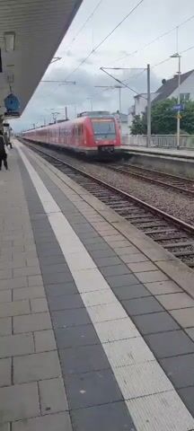 A red train approaches a station platform. The train is long and appears to be traveling at a moderate speed. The platform is paved with gray and white tiles. A single person is standing on the platform, looking towards the approaching train. Overhead wires are visible, suggesting the train is electric. Buildings and trees are visible in the background. The sky is overcast.