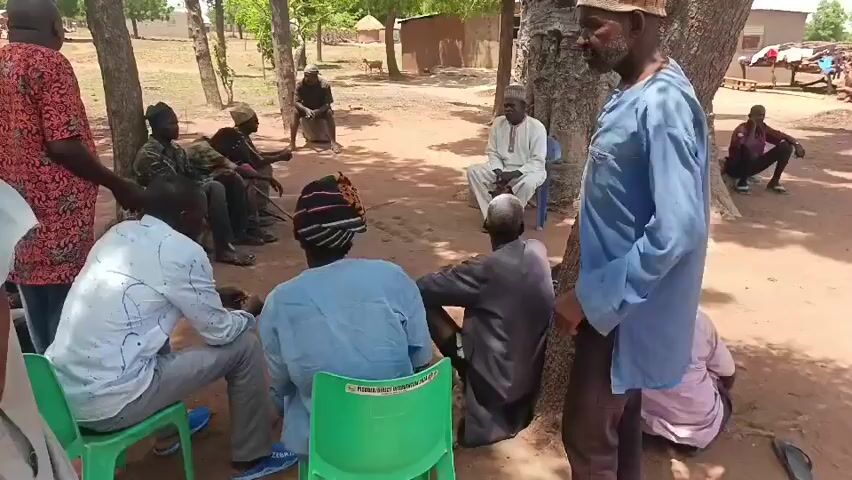 A group of approximately 15 men are gathered in a circle on a dirt ground under trees.  Some men are seated in green plastic chairs, some are sitting on the ground, and some are standing. One man in a red and orange patterned shirt is standing and appears to be addressing the group, gesturing with his hands.  Another man stands near a tree wearing a blue shirt. In the background are simple buildings and more trees. The scene appears to be outdoors in a rural area.