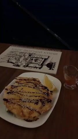 A close-up shot focuses on a white plate with a food item drizzled with dark sauce and sprinkled with sesame seeds. A slice of lemon sits on the side of the plate. Next to the plate is a small glass dish containing a reddish sauce. A menu with a drawing and text in Russian is positioned behind the plate. The scene is set on a wooden table. In the background, a body of water is visible under a dark, cloudy sky, with distant lights on the horizon. The camera angle slightly shifts, revealing more of the water and the darkening sky as the video progresses.