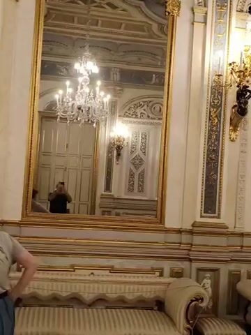 The video opens with a close-up of ornate wall decorations, gold trim, and a large mirror reflecting a chandelier and sconce. A section of a couch with tufted upholstery is visible in the foreground. The camera pans left, revealing more of the richly decorated room, including a woman in a black top standing near a wall. The view then shifts to a large floral arrangement centerpiece with palm fronds and red accents, surrounded by people who appear to be tourists. The camera moves again, showing more of the elaborate ceiling and walls, another woman looking up, and a group of people on the left side of the frame. The scene is an indoor setting, likely a palace or grand hall, characterized by opulent decor.