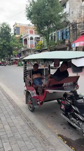 The video shows a red and silver tuk-tuk vehicle parked on the side of a street. Two men are seated in the passenger area of the tuk-tuk. The man on the left is wearing a dark shirt and blue jeans, and he is smiling and waving at the camera. The man on the right is wearing a dark long-sleeved shirt and jeans and is also smiling. A motorcycle is parked behind the tuk-tuk. The street appears to be in an urban area with buildings and trees visible in the background. The road is paved, and there is a brick sidewalk in the foreground. The sky is overcast. The camera is moving slightly, giving a dynamic perspective of the scene.