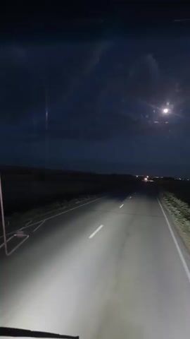 This media file depicts a nighttime drive on a highway in Baiqadam, Kazakhstan. The scene is illuminated by the vehicle's headlights, casting a bright glow on the asphalt road and the surrounding sparse vegetation. The sky is dark, with clouds partially obscuring a bright full moon that shines in the upper right quadrant of the frame. In the distance, a faint line of lights suggests a town or city.

The perspective is from inside the vehicle, looking forward. We see parts of the vehicle's dashboard and windshield wipers in the foreground, hinting at the point of view. The road has white lane markings, and there are reflective markers on the side of the road. The overall mood is quiet and perhaps a bit isolating, characteristic of a nighttime journey through a less populated area. The time of day is clearly night, and the weather appears clear enough for the moon to be visible, though some clouds are present. There are no people visible in the media file. The activity taking place is driving on a highway. There is no visible text.