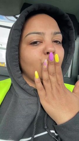 A woman wearing a dark gray hoodie and a neon yellow safety vest is sitting in a vehicle. She holds up her hand, showing off her nails, which are painted yellow and purple with a marbled design on one. She pouts and blows a kiss towards the camera. The interior of the vehicle is visible in the background.
