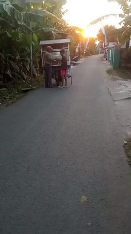 A young person wearing a dark t-shirt and pink shorts walks away from a food cart on a paved road at sunset. The food cart is being pushed by an adult wearing a red cap and a light-colored shirt. The food cart is a mobile unit with a canopy, and it appears to be on a stand or a small vehicle. Banana trees and other foliage line the left side of the road, and houses are visible on the right. The sun is setting in the distance, casting a warm glow on the scene.