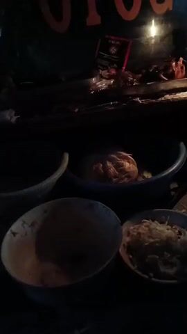 The video shows a dimly lit scene, possibly a food stall or preparation area at night. A single lit candle provides the primary light source, casting long shadows and illuminating a few select items. In the foreground, there are several bowls. One bowl on the left appears to be mostly empty with some residue at the bottom. To the right of that, there is another bowl filled with shredded white material, possibly cheese or noodles. Further to the right, another bowl contains what looks like noodles. In the middle ground, a blue plastic basin holds a large, round object wrapped in what appears to be plastic or cloth. Behind this, on a surface that might be a counter, a few more bowls are stacked, and some food items, possibly meat, are visible. A red and black packet or label is also present on this surface. The background is dark, but a portion of an orange sign with dark lettering is visible at the top. The overall impression is one of simple, perhaps impromptu, food preparation or service in low-light conditions.