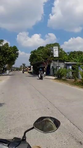 The video appears to be captured from the perspective of someone riding a motorcycle or scooter on a paved road. The rider's left side mirror is prominently visible in the foreground, reflecting the sky. The road stretches ahead, with buildings and trees lining both sides. Several other motorcycles are visible on the road, some moving towards the camera and others away. The sky is blue with scattered white clouds, suggesting fair weather. The buildings on the right appear to be commercial establishments, with some signs visible. The overall scene depicts a daytime view of a street or road in what appears to be a populated area, possibly a town or suburban environment.