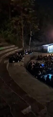 A nighttime outdoor amphitheater scene. A crowd of people are seated on the tiered seating and on the ground in front of a stage. Many of the people are holding up their phones, illuminated by their screens. The stage is lit, with a large screen at the back, and there are structures on the stage that appear to be part of a performance or display. A streetlamp is visible in the upper right of the frame, providing additional light. The overall atmosphere suggests an outdoor event or gathering at night.
