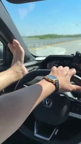 The video shows a person driving a car on a highway during a sunny day.  The driver is wearing a watch on their left wrist and has their bare feet up on the dashboard.  The speedometer reads 139.  Other cars are visible on the highway in the distance. The driver is holding the steering wheel with their right hand.