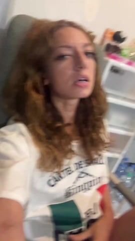 A woman with long, wavy auburn hair sits in an office chair wearing a white t-shirt that reads "Cartel de Sinaloa Kingpin El." Her lips are slightly parted and she has a playful expression. The camera is held in her hand, creating a selfie-style shot. The background includes a white storage unit with various objects, including clear plastic containers, bottles, and other items. There is also a glimpse of a gray wall and possibly another piece of furniture. The lighting appears to be indoor lighting, possibly fluorescent. The woman moves the camera around slightly as she sits in the chair.