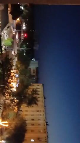 This video appears to be taken from a high vantage point, possibly a window or balcony, overlooking a cityscape at dusk or early evening. The scene is dominated by a tall, light-colored apartment building with numerous windows, some of which are illuminated, suggesting occupancy. Trees with dark foliage are visible in the foreground, partially obscuring the view of other buildings and structures. The sky is a deep blue, transitioning to a lighter shade towards the horizon, typical of twilight. There are some lights visible in the distance, possibly streetlights or lights from other buildings, indicating an urban environment. The camera is positioned such that the buildings are viewed from a side angle, and the overall perspective is tilted. The resolution is somewhat low, and the image quality is further degraded by motion blur, suggesting the video was recorded handheld or while moving. No people are clearly discernible, and the focus is on the architectural elements and the transition from day to night. The overall mood is quiet and urban, with the illuminated windows offering a sense of life within the otherwise darkening landscape. There is no visible text in the video. Given the location context of Qorowul, Uzbekistan, the scene likely depicts a residential area in that region during the evening hours.