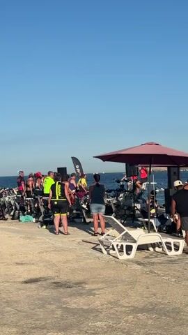 A group of people stand around a row of stationary bicycles parked on a paved outdoor area. Some people are wearing athletic clothing and racing numbers, suggesting a sporting event. The bicycles are lined up facing a body of water, presumably the sea, where boats can be seen in the distance. The sky is clear and blue, indicating a sunny day. There are flags and lounge chairs present, adding to the event atmosphere. Some individuals are interacting with each other or adjusting their equipment.