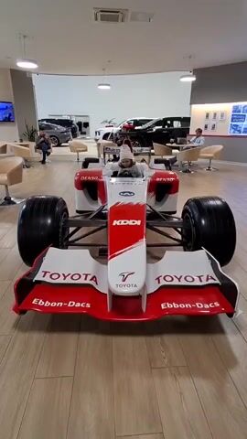 A child wearing a striped beanie is sitting in the cockpit of a red and white Formula 1 race car on display in what appears to be a car dealership or showroom. The child is holding the steering wheel and looking forward. The car has various sponsor logos, including "TOYOTA," "KDDI," "DENSO," and "Panasonic." In the background, other cars, seating areas, and display walls are visible. The lighting suggests an indoor environment. The camera perspective is from the front of the race car, looking towards the child.