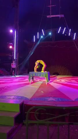 A woman in a yellow outfit is performing a backbend on a small table in a circus tent. The tent floor is decorated with red, white, and yellow stripes and blue stars. A man in a white shirt and black pants stands to the side, holding a microphone and watching the performance. The tent is lit with bright spotlights and string lights. There is a metal structure above the stage, and red curtains behind the performer.