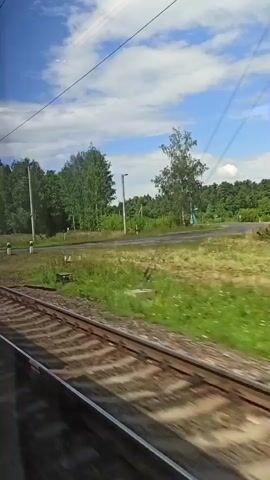 This is a video of a train moving along a track. The camera is looking out the window of the train and shows the scenery passing by. The scenery consists of green fields, trees, and a blue sky with white clouds. There are also power lines and poles visible along the side of the tracks. The train tracks are made of wood and metal and are visible in the foreground. The motion of the train causes the landscape to appear blurred, creating a sense of speed.