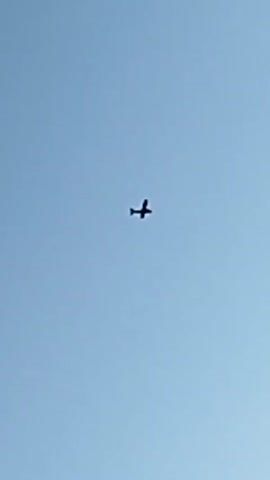 A small airplane is seen flying against a clear blue sky. The plane is silhouetted against the sky, making it difficult to discern any specific details. The perspective is from below, looking up at the aircraft as it moves across the frame. The video appears to be shaky, suggesting it might have been recorded handheld.
