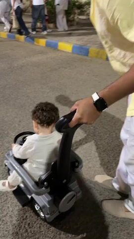 A person is pushing a child in a toy car with a stroller handle. The child is seated facing away from the viewer. The person pushing the car is wearing a yellow shirt and white pants, and a watch on their left wrist. The child is wearing a light-colored outfit. The scene appears to be an outdoor walkway or paved area, possibly a park or promenade, with trees and streetlights visible in the background. Other people are also present in the background, walking or standing. The lighting suggests it is evening or night.