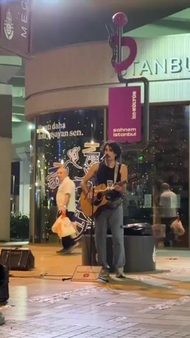 A man stands on a paved walkway playing an acoustic guitar and singing. He is wearing a black tank top, grey pants, and white sneakers. A microphone stand is positioned in front of him. To his left, a person walks by carrying a white plastic bag. Behind the man, a building has large windows displaying graphics and text, including the word "ISTANBUL." A purple banner hangs from a pole in front of the building, displaying the text "İBB KÜLTÜR SAHNEM İSTANBUL." The scene appears to be outdoors at night, with artificial lighting illuminating the area.