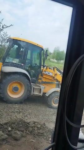 This video shows a yellow JCB backhoe loader parked on muddy ground, with trees and foliage in the background under a cloudy sky. The perspective appears to be from inside another vehicle, looking out through a window. The backhoe has its front loader raised. Visible text on the backhoe includes "JCB" and a license plate that reads "10487 T AH 30X". The scene suggests an outdoor work site, possibly under construction or undergoing excavation. The weather appears overcast. The time of day is not definitively indicated but the lighting suggests daytime.