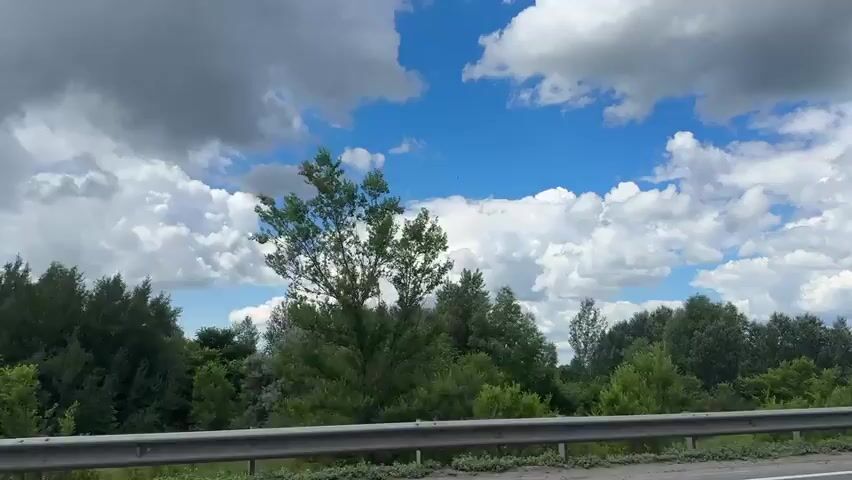 The video shows a view from a vehicle moving on a road with a gray metal barrier in the foreground. On the left side of the road, there is a dense line of green trees. The sky is blue with scattered white and gray clouds. A gray SUV passes by on the road. Further in the distance, more trees and green fields can be seen. A large gray semi-trailer truck drives past on the right. The camera seems to be mounted on the side of the moving vehicle.