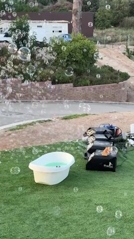 The video shows a backyard scene with a white baby bathtub filled with green liquid. Next to the bathtub is a black bubble machine, which is producing a large quantity of bubbles. The bubbles are floating in the air and filling the frame. The background includes a brick wall, trees, a house, and a car. The ground is covered with artificial grass.