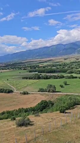 The video pans across a rural landscape under a partly cloudy blue sky. In the foreground, a dry, grassy hill is bordered by a wire fence with wooden posts. Beyond the hill, a patchwork of green fields and trees stretches into the middle ground. Further in the distance, a dense village with red-tiled roofs is nestled among the trees. In the background, a range of mountains forms the horizon. There are no people or animals visible in the video.