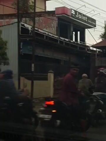 The video shows a busy street scene, likely in Southeast Asia, with numerous motorcycles and a few cars. The individuals are predominantly male and are seen riding motorcycles, many wearing helmets. The environment appears to be urban, with buildings and a gas station visible in the background. The gas station has the prominent "PERTAMINA" logo. There are signs that appear to be in a local language. The lighting suggests it is daytime, possibly overcast. The overall impression is of a traffic situation, with vehicles moving slowly or stopped. The camera is positioned inside a vehicle, providing a view through the windshield or windows.
