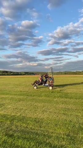 Two people are in a powered paraglider, which is on the ground on a grassy field. The paraglider has a large wing that is beginning to inflate with the wind. The sky is blue with white clouds, and trees and buildings are visible in the distance.