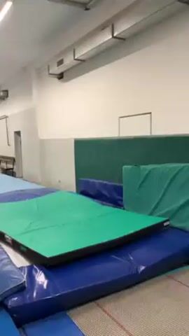 A young man in a black t-shirt and red shorts performs a handspring on a green square gym mat on top of a blue padded surface. The man lands on his feet in the center of the mat, facing the camera. The gym has white walls, a blue padded floor, and a green padded section against the wall.