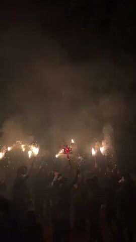 A nighttime outdoor scene shows a crowd of people holding up flaming torches and other glowing objects. Sparks are flying from the torches and creating a smoky atmosphere. The crowd appears to be celebrating an event or festival. Some people are raising their arms in the air.