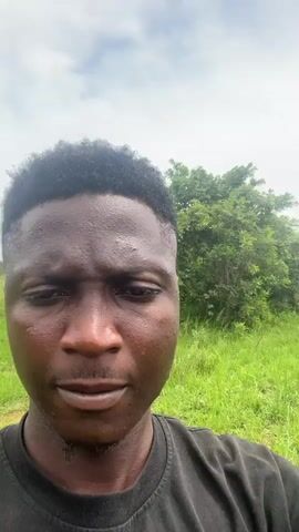 A young black man wearing a black t-shirt with a colorful graphic on the front is filming himself with a phone, holding it in his right hand and giving a thumbs up. He is outdoors in a grassy field with trees in the background. The sky is overcast.
