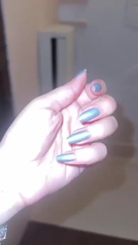 A close-up shot shows a left hand with long, coffin-shaped nails painted a metallic, shimmery light blue-gray color. The hand is positioned with the palm facing the camera, and the fingers are slightly curled. The background is out of focus, but appears to be an indoor setting with neutral-colored walls and some indistinct furniture or fixtures. The lighting suggests an indoor environment, possibly artificial light. The video loops.