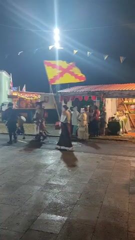 The video shows a nighttime scene at what appears to be a historical reenactment or festival. A group of people dressed in historical costumes, possibly medieval or Renaissance, are walking across a paved area. Some of them are wearing armor and carrying swords. In the background, there are stalls or booths with awnings and decorations, and string lights are visible overhead. A flag with a yellow and red cross pattern is flying from a pole. The ground is wet, suggesting recent rain. The overall atmosphere is festive and atmospheric.