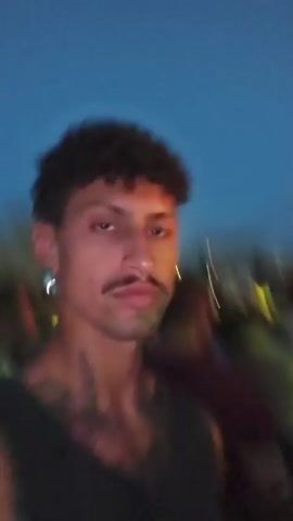 A first-person perspective video shows a man with curly hair and a mustache at an outdoor concert. He is wearing a dark tank top with tattoos on his arm. Behind him, a crowd of people is visible, many wearing bright neon green shirts. A stage with bright yellow and green lights is in the background, with a DJ booth and a DJ present. The sky is dark, indicating it is evening or night. The man waves at the camera.