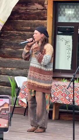 In this media file, a young person with long, light brown hair, wearing a black beret, a patterned knit sweater in shades of brown, beige, and grey, and loose-fitting brown pants, stands on a wooden stage. They are holding a microphone and appear to be singing or speaking, with one hand near their mouth and the other gesturing towards their head. A red and green plaid shirt is tied around their waist.

Behind the performer, a rustic wooden building serves as the backdrop. A window with a white, decorative curtain is visible. To the left of the performer, a white folding chair and some green plants are present. In front of the building, a table is covered with a vibrantly patterned tablecloth featuring diamond shapes in red, orange, yellow, and white.

To the left of the frame, a smartphone is being held, recording the scene. A microphone stand is also visible on the right side of the stage. The lighting suggests it could be daytime, possibly late afternoon or early evening, with diffused light. The overall atmosphere appears to be a performance or gathering in an outdoor, possibly rural setting, indicated by the wooden structures and natural elements. The setting is identified as Старые Быги, Russia.