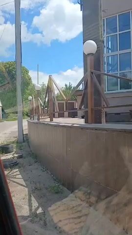 The video captures a street scene on a cloudy day. In the foreground, a weathered concrete wall runs horizontally, with tires and some debris scattered on the ground to its left. Above the wall, a row of wooden structures with triangular roof supports and white globe lights are visible. Beyond these structures, several cars are parked, including what appears to be a police car with flashing lights. Further in the background, a light-blue building with a steeple, possibly a church, stands against the sky. The overall environment suggests an outdoor area with some commercial or institutional buildings.