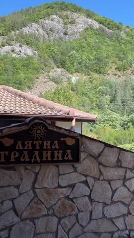 The video shows a stone wall with a wooden sign in front of a building with a tiled roof. The building is nestled against a lush green mountainside. The sky is a clear blue. The sign has a sun emblem and text written in Cyrillic script. The camera pans slightly to the right, revealing more of the vegetated mountain slope.