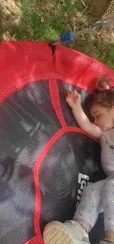 The video shows a young child, appearing to be a toddler, on a trampoline outdoors. The child is wearing a light-colored t-shirt with a cartoon animal graphic and light-colored pants with a patterned design, along with white sneakers. The trampoline has a red safety pad around the edge and a black bouncing surface. The child is seen lying down and then rolling over, seemingly enjoying themselves. The setting appears to be a grassy area, suggesting a backyard or park. The lighting indicates it is daytime, possibly sunny. The video is filmed from a high angle, looking down onto the trampoline. No text is clearly visible.