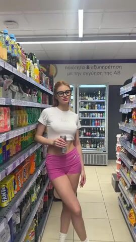 A young woman with red hair and glasses stands in a brightly lit supermarket beverage aisle in Paleo Faliro, Greece, during what appears to be daytime. She is wearing a white cropped t-shirt with "Juicy" written on it, pink shorts, and has a belly button piercing. She holds a metallic can, rotating it as if presenting it to the camera, with the word "Juicy" also visible on the can.

The aisle is lined with shelves stocked with various bottled and canned beverages, including sodas, juices, and energy drinks. Behind her, a glass-door refrigerator unit is also filled with drinks. Above the refrigerator, a sign on the wall displays Greek text: "ποιότητα & εξυπηρέτηση με την εγγύηση της AB," which translates to "quality & service with the AB guarantee." This text identifies the location as an AB Vassilopoulos supermarket. The woman appears calm, looking directly at the camera while holding the can.