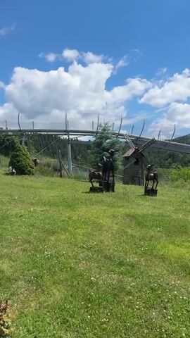 The video pans across a grassy hillside on a sunny day. In the foreground, there are several decorative elements including two metal horse statues, a small wooden windmill, and three planters with greenery and flowers. Further in the background, a raised track, likely for a rollercoaster or alpine slide, winds its way up the forested hillside. The sky is a vibrant blue with fluffy white clouds. The overall impression is a scenic outdoor recreational area.