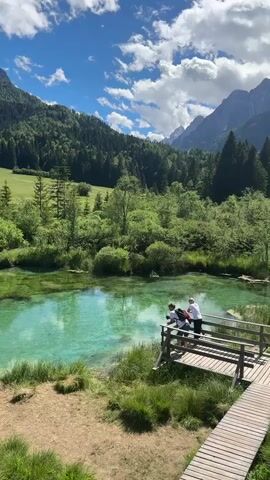 The video shows a tranquil natural scene with a crystal-clear turquoise lake. A wooden boardwalk leads to a viewing platform overlooking the lake. On the platform, two individuals, appearing to be young people, are observing the water. The surrounding environment is lush and green, with dense forests and mountains in the background under a bright, cloudy sky. Another person is visible further down the boardwalk.