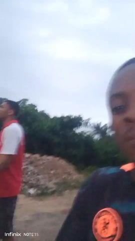 The video appears to be a selfie video taken outdoors in Atani, Nigeria, likely during the daytime under cloudy or overcast skies. The footage is shaky and motion-blurred, suggesting the person recording is in motion, possibly walking or running.

The primary subject is a young Black person, who is the one recording the video. They are wearing a dark t-shirt with some colorful, possibly orange and blue, accents on the collar and shoulders. There's a circular emblem on the left side of the shirt, which resembles a sports team logo, specifically the Chelsea FC crest.

In the background, glimpses of other people are visible, also in motion and blurred. One person is seen wearing a red and white shirt and dark shorts. The setting appears to be somewhat rural or undeveloped, with some foliage and rough terrain visible. The ground seems to be made of dirt or gravel.

The overall mood is informal and energetic due to the movement and the selfie format. The cloudy weather might indicate a humid or potentially rainy climate, common in Nigeria. The "Infinix NOTE 11i" watermark in the bottom left corner of the screen confirms the device used to capture the video. The audio, though not described, would likely capture ambient sounds of the environment and any conversation or exclamations from the individuals.