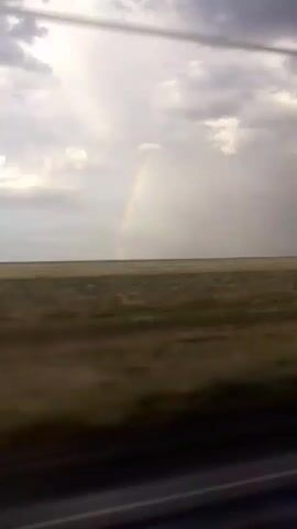 The video shows a landscape with a faint rainbow in the sky above a flat, grassy plain. The image appears to be captured from a moving vehicle, as indicated by the motion blur in the foreground and midground. The sky is overcast with clouds, and sunlight is breaking through in some areas, creating a dramatic lighting effect. The overall scene suggests a natural environment, possibly a prairie or desert, under a moody sky.