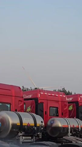 A row of red semi-trucks with silver tankers are parked outside under a hazy, light blue sky. A thin, white pole with a red tip extends vertically from the top of one of the truck cabs and a light-colored object is seen extending from the top of the pole. The pole appears to be moving, with the object at the top of it moving in a sweeping motion. It is unclear what the pole is attached to or what its purpose is. The trucks appear to be new, with some still having protective coverings on their mirrors.