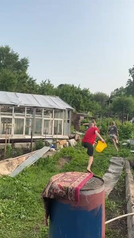 The video shows a backyard with a greenhouse on the left and overgrown vegetation. In the center background, a person in a red shirt and dark shorts runs from right to left, carrying a yellow bucket. Behind them, another person in a light-colored shirt and dark shorts follows. In the foreground, a large blue and red barrel is visible, draped with a colorful rug. The ground is covered in grass and dirt paths. The sky is visible and appears hazy.