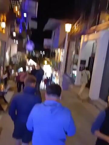 The video shows a nighttime street scene in a tourist area, likely a village or town. People are walking along a narrow pedestrian street lined with shops and restaurants. The street is illuminated by decorative string lights and the lights emanating from the businesses. Many of the restaurants have outdoor seating, with people sitting at tables. The camera appears to be held by someone walking down the street, capturing the scene as they move.