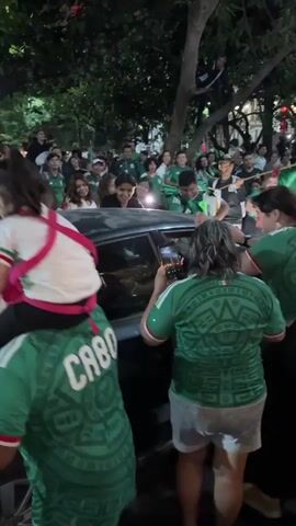 A group of people, most wearing green shirts with the Mexican flag on them, are gathered around a dark colored car on a street at night. Many are holding up Mexican flags. One person is sitting on the shoulders of another person, who is wearing a green shirt that says "CABO" on the back. Some people are leaning on the car, and others are holding up phones to take pictures or videos. There are trees and lights visible in the background. The overall atmosphere appears to be celebratory.