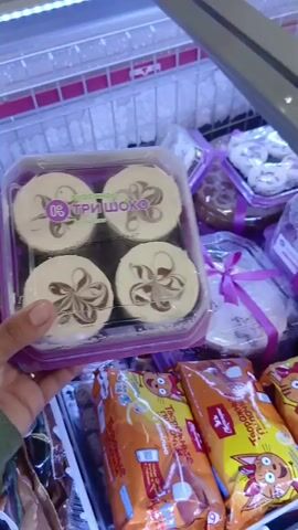 The video shows a person's hand holding a clear plastic container with four round desserts inside. The desserts have a swirl pattern of dark chocolate on a light-colored base. The container is purple. In the background, there are other desserts in clear plastic packaging, many tied with purple ribbons. The desserts appear to be in a refrigerated display case in a store. There are also some packaged food items at the bottom of the frame with orange and yellow packaging featuring cartoon characters.