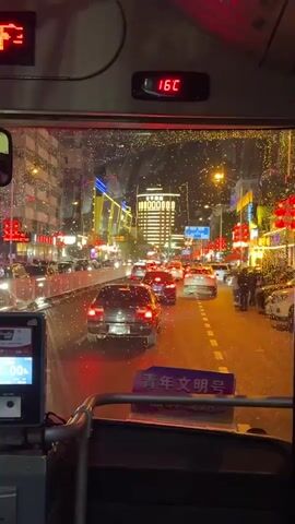 From inside a bus, facing forward through the front window, a cityscape is visible at night. It is raining or snowing lightly, with droplets and specks on the glass. The street below is filled with cars and pedestrians, their forms blurred by movement and the weather. The buildings lining the street are brightly lit with colorful neon lights and strings of lights, contributing to the festive atmosphere. A digital display above the window shows a temperature of "16C", which changes to 19:52 and a text prompt. The camera is shaky, indicating movement, and a sign that reads "青年文明号" is visible below the windshield.